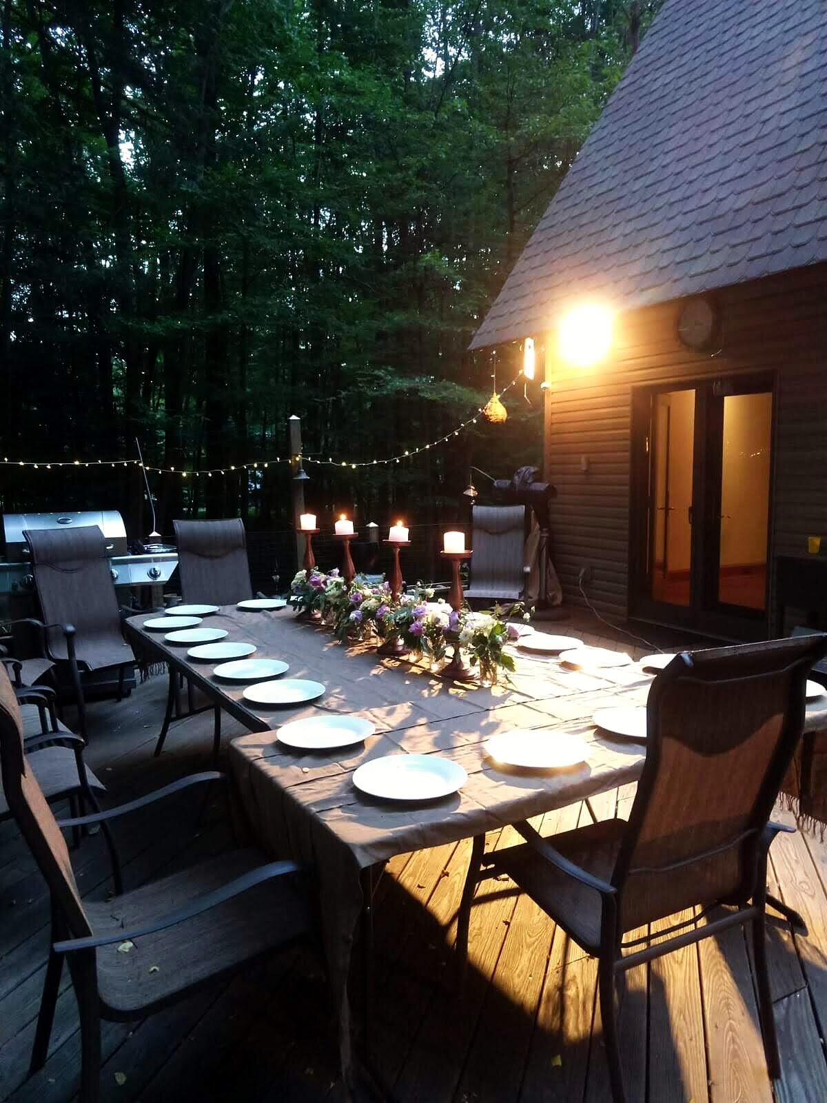 The deck at dusk: a long table set for dinner under string lights, the house glowing behind.