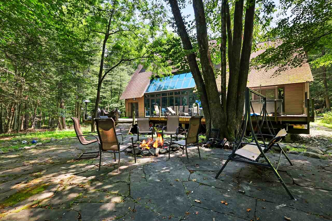 The back of the house at dusk: the greenhouse window wall lit from within, a flagstone patio, a fire pit, and tall forest trees.