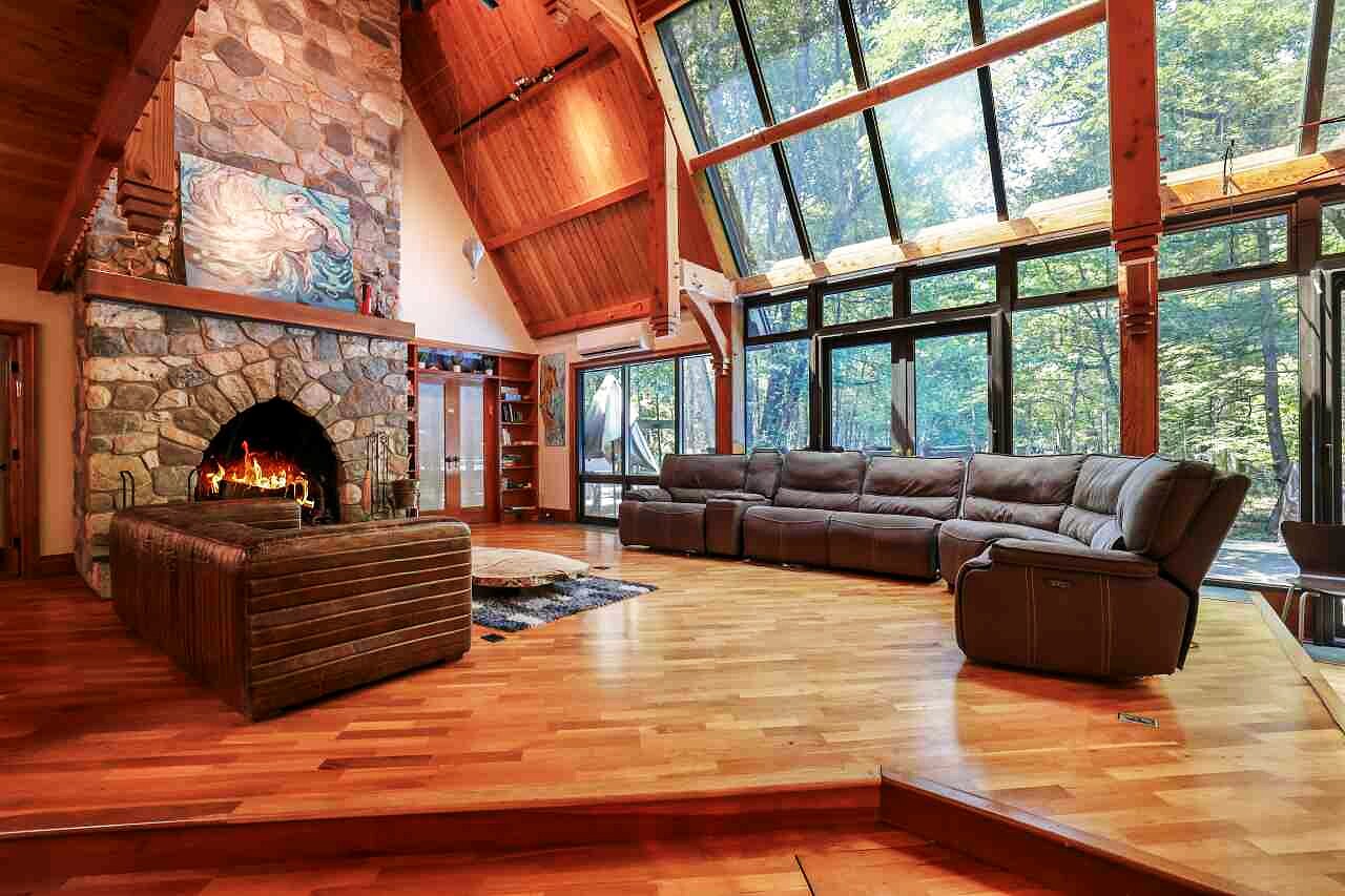 The grand salon: stone fireplace, sectional seating, and the two-story greenhouse window wall opening to the forest.